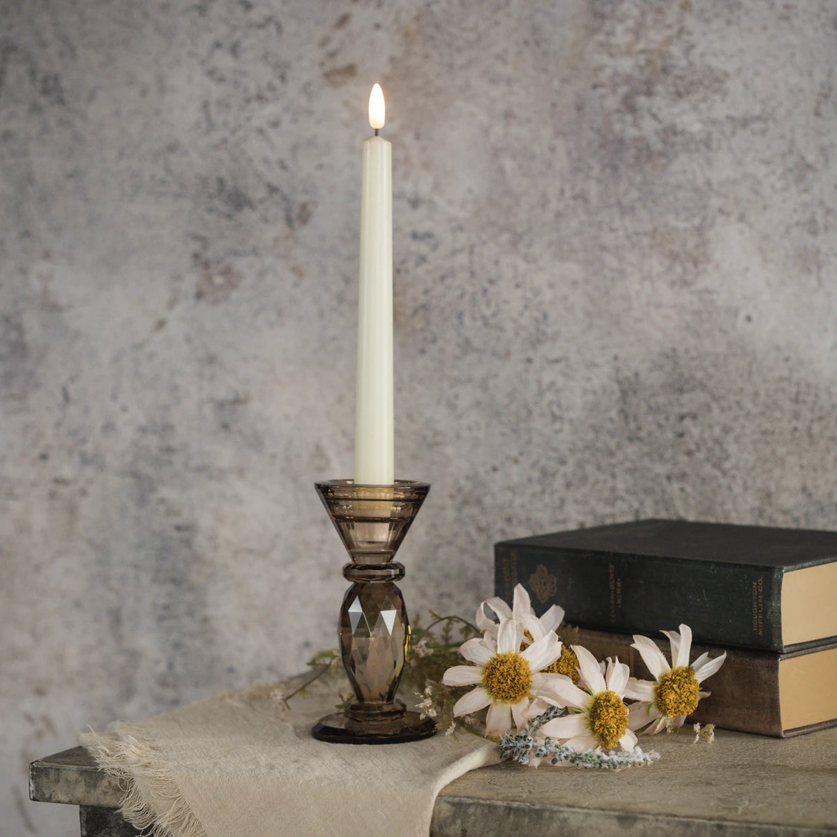 A lit white taper candle in a dark amber-brown faceted glass holder, styled on a rustic table with books and white daisies against a textured gray wall.
