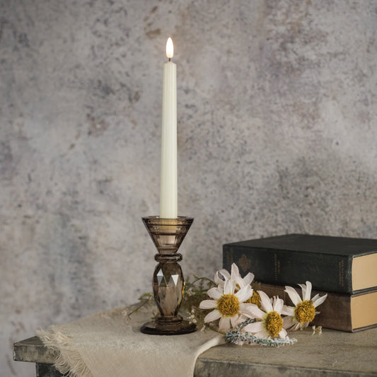 A lit white taper candle in a dark amber-brown faceted glass holder, styled on a rustic table with books and white daisies against a textured gray wall.