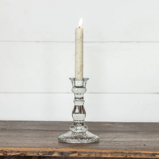 A lit taper candle in a clear glass candlestick holder with a speckled cream wax, set on a rustic wooden surface against a white paneled wall.