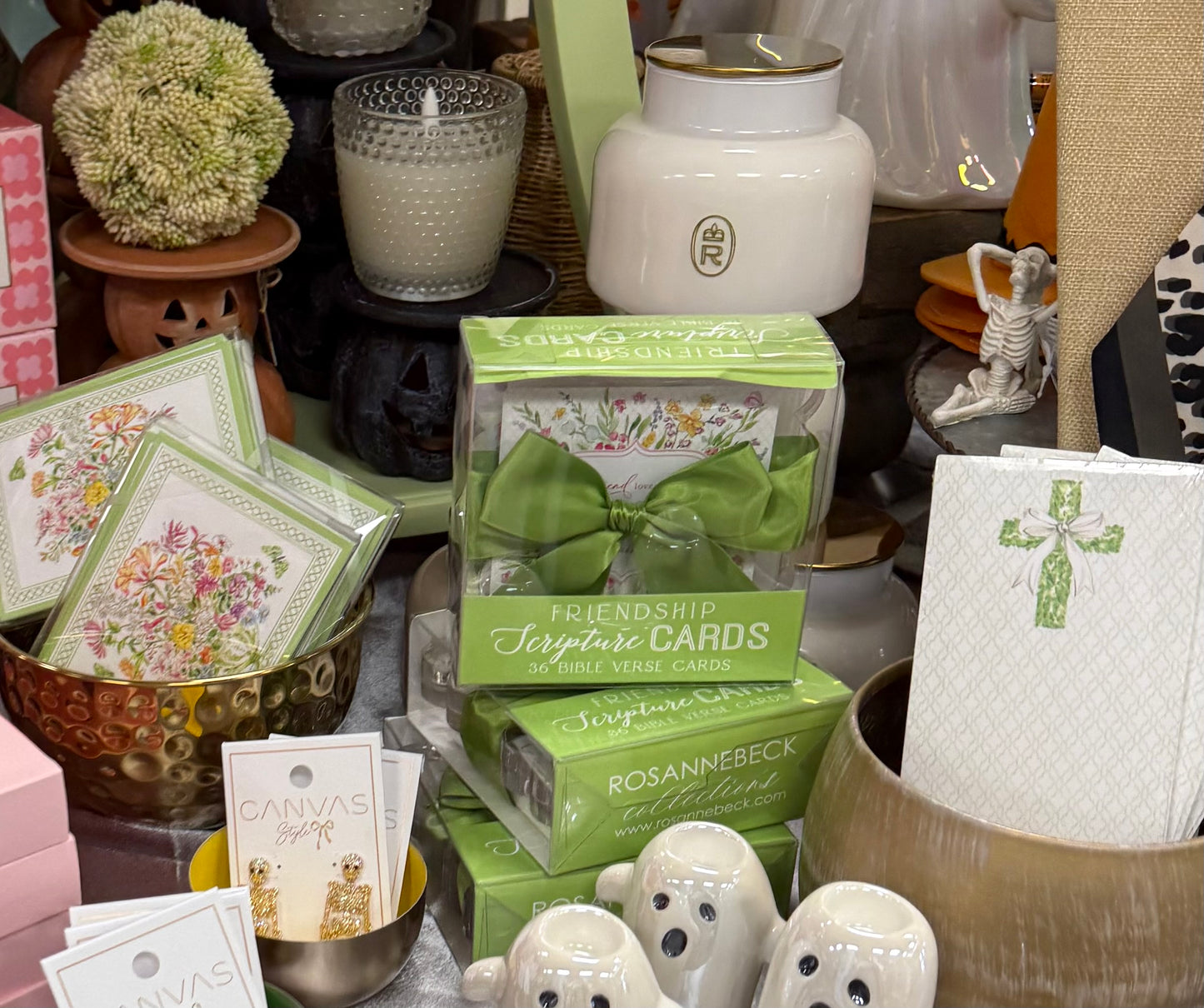 A styled tabletop display featuring multiple boxed and packaged decorative cards and small ceramic figurines. Prominent products include green-labeled card boxes reading 'Friendship Jumbo Cards' and 'Rosanne Beck' cards, floral card designs in a bowl, and white ceramic ghost and skeleton figures.