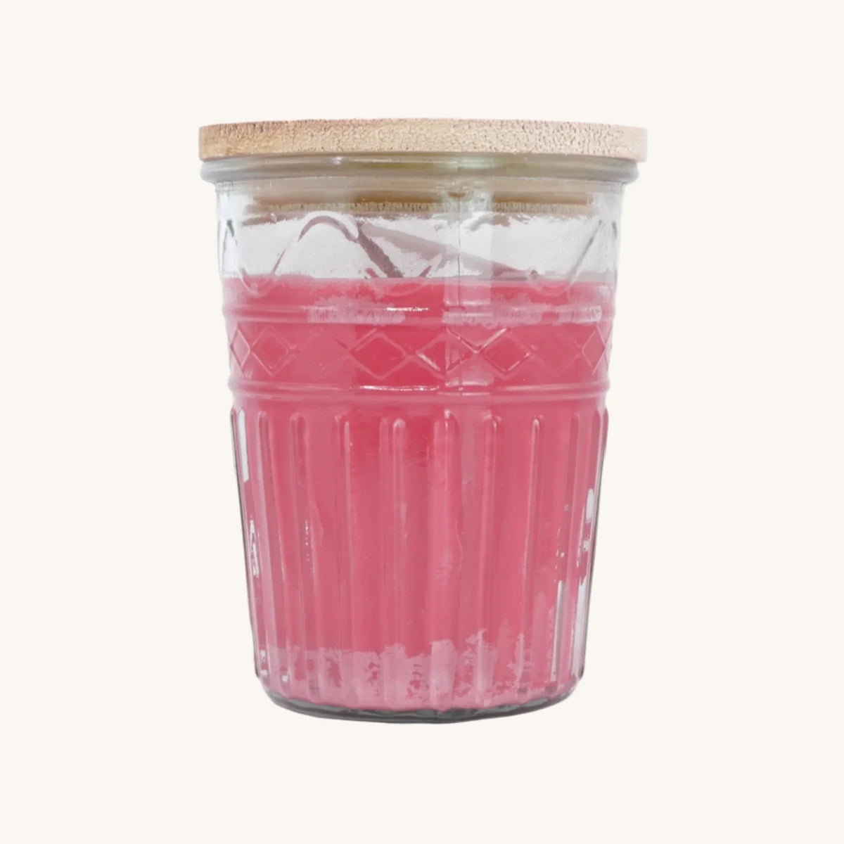 A pink wax candle in a clear ribbed glass jar with a light natural wood lid, set against a plain off-white background.