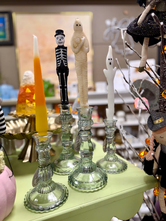 Halloween-themed glass candlesticks on a pale green table, featuring a skeleton figure, a mummy, a ghost, and a white ghost candle, with other seasonal decor blurred in the background.