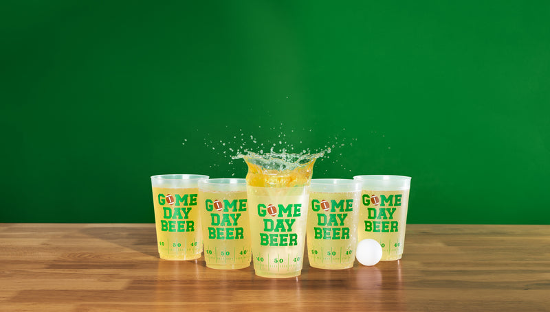 Five clear plastic cups with green and yellow 'GOME DAY BEER' text are lined up on a wooden table against a solid green background. A splash of yellow liquid is captured in the center cup, with a white ping-pong ball resting beside it.