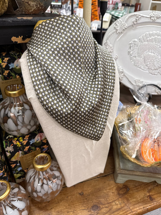 A folded patterned bandana or scarf displayed on a beige fabric stand on a wooden shelf, surrounded by home decor items including glass bottles, a white decorative plate, and wrapped orange pumpkins.
