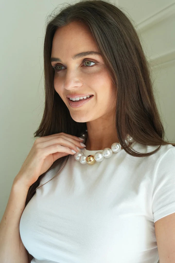 A woman wearing a white crew-neck t-shirt and a large beaded gumball pearl necklace with a single gold bead accent, posing indoors against a light beige wall.