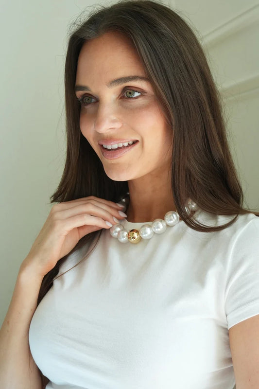 A woman wearing a white crew-neck t-shirt and a large beaded gumball pearl necklace with a single gold bead accent, posing indoors against a light beige wall.