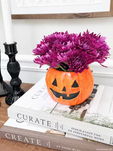 An orange jack-o'-lantern vase filled with bright purple flowers, displayed on stacked books on a tabletop with a black candlestick and white wall in the background.