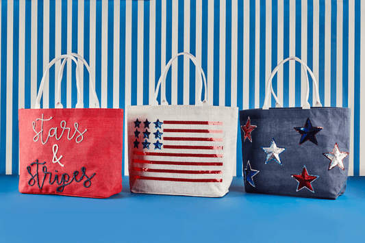Three fabric tote bags with white handles are lined up against a blue and white striped background. The left bag is coral red with white script text reading 'Stars & Stripes'.