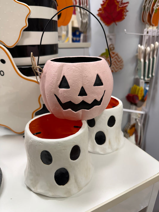 A pink jack-o'-lantern candy bucket with a black handle, displayed on top of two white ghost-shaped buckets with black eye cutouts, on a white table in a store setting.