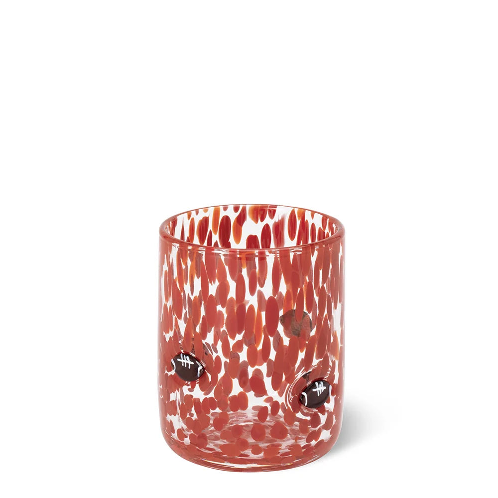 A clear glass tumbler with a vibrant red and white speckled pattern and two small black football charms attached to the sides, shown on a plain white background.
