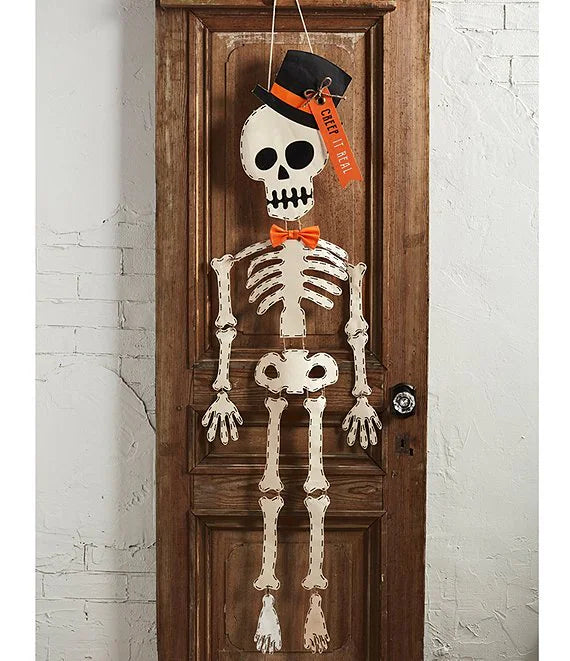 A hanging Halloween skeleton decoration with a black top hat, orange bow tie, and orange hanging tag, displayed on a brown wooden door against a white brick wall.