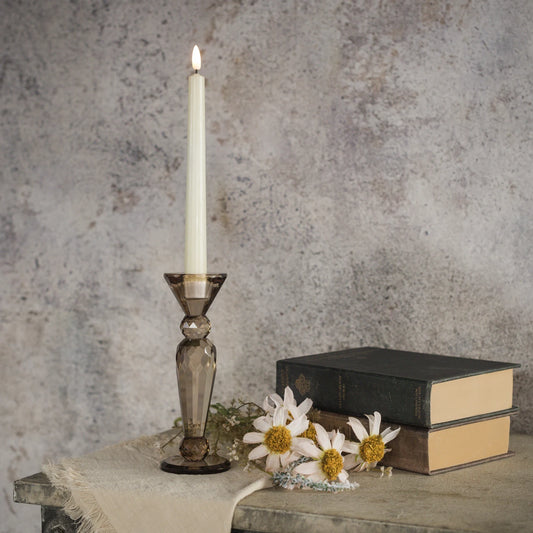 A tall white taper candle in a smoky amber glass candlestick holder, styled on a rustic table with vintage books and white daisies against a textured gray wall.
