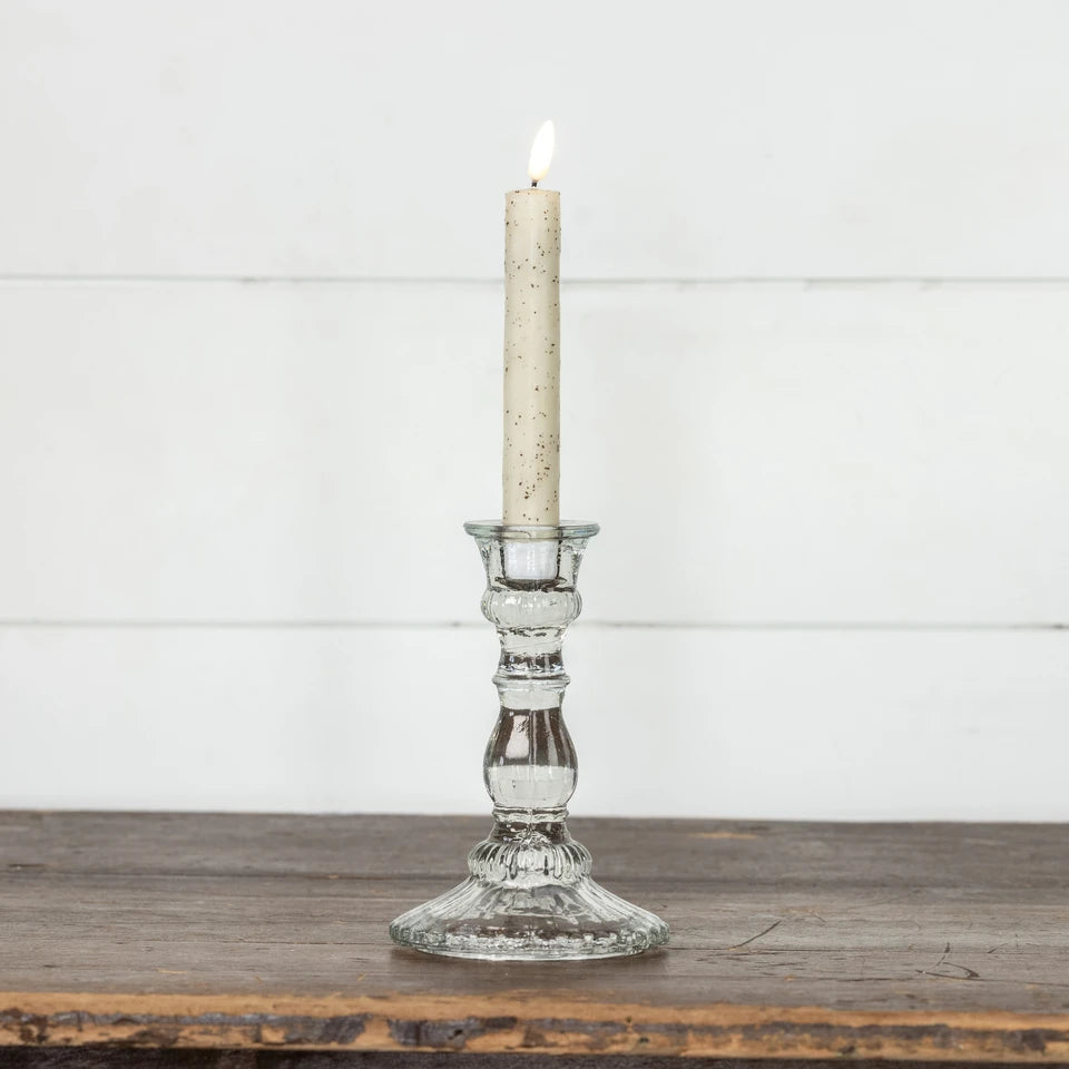 A lit taper candle in a clear glass candlestick holder with a speckled cream wax, set on a rustic wooden surface against a white paneled wall.