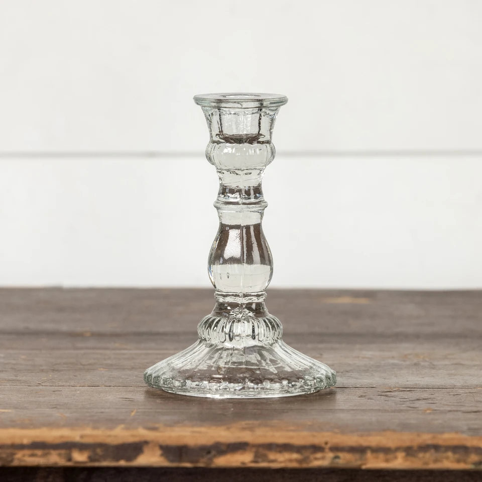 A clear glass candlestick holder with a fluted base and a ribbed stem, standing on a rustic wooden surface against a white paneled wall.