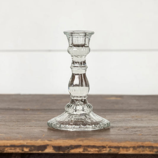 A clear glass candlestick holder with a fluted base and a ribbed stem, standing on a rustic wooden surface against a white paneled wall.