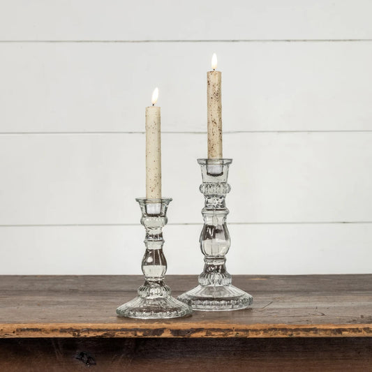 Two clear glass candlestick holders with lit taper candles, displayed on a rustic wooden surface against a white shiplap wall.