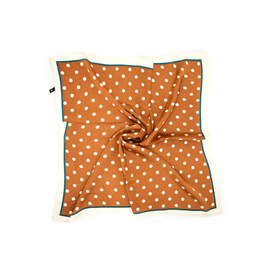 A square scarf with a rust-orange background, white polka dots, and a thin dark teal border, styled in a central swirl on a solid white background.