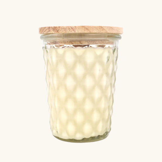 A clear glass jar candle with a textured honeycomb pattern, filled with creamy off-white wax and topped with a light natural wood lid, shown against a solid off-white background.