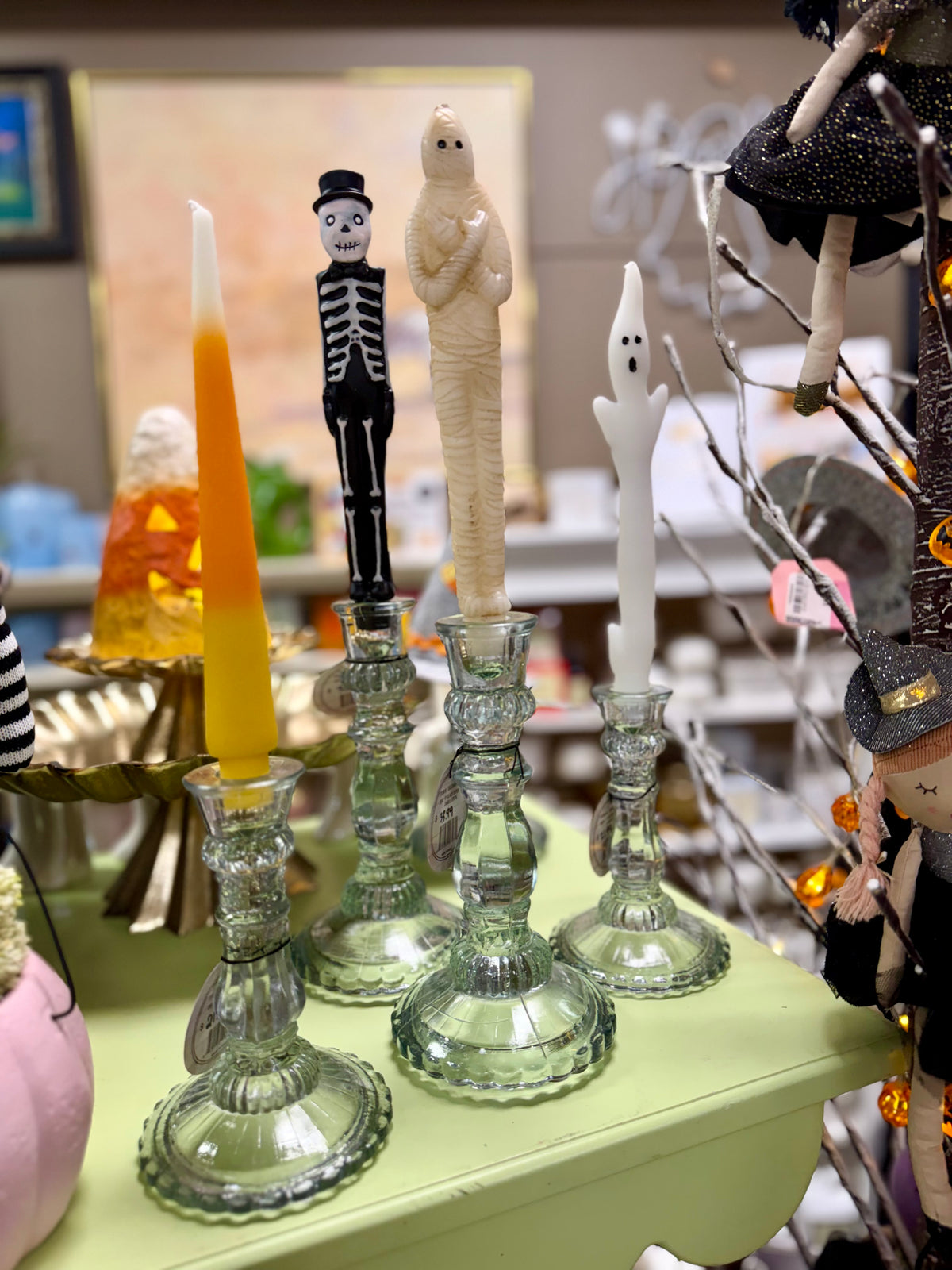 Halloween-themed glass candlesticks on a pale green table, featuring a skeleton figure, a mummy, a ghost, and a white ghost candle, with other seasonal decor blurred in the background.