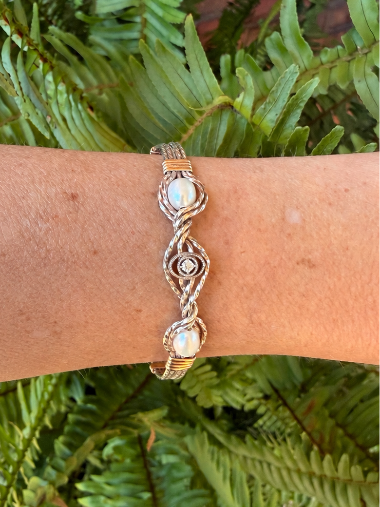 A close-up of a person's wrist wearing a handmade 14k gold and sterling silver metal wire bracelet with two white pearl like stones and a small central clear stone, set against a background of green fern leaves.