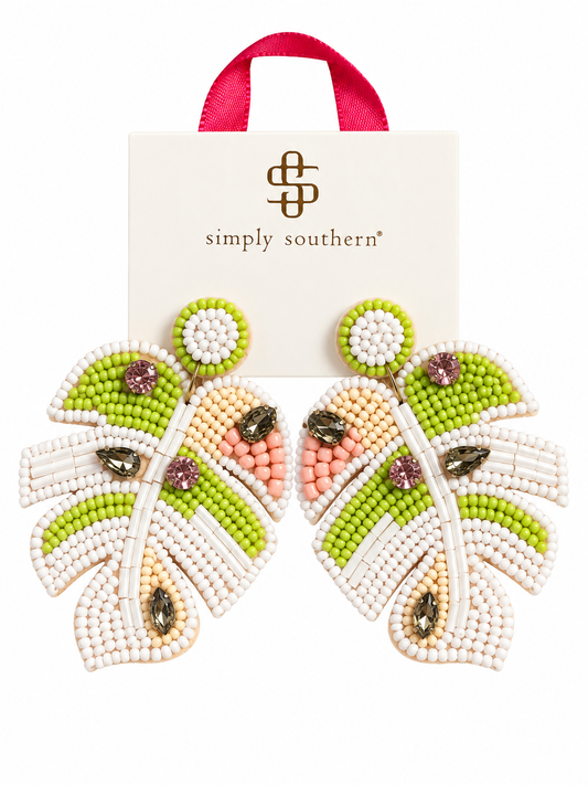 A pair of beaded leaf-shaped earrings by Simply Southern, featuring green, cream, and peach seed beads with dark brown rhinestones, displayed in front of a cream-colored branded card with a pink ribbon handle.