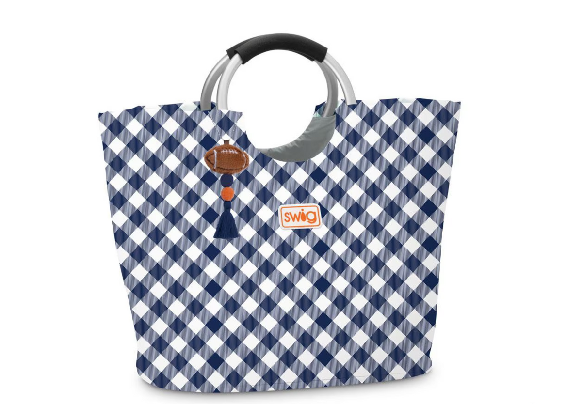 A large tote bag with a navy blue and white gingham checkered pattern, featuring a football-shaped charm with a tassel and a small orange and white Swig logo patch. It has silver-tone metal handles with black grips, shown on a plain white background.  Auburn.