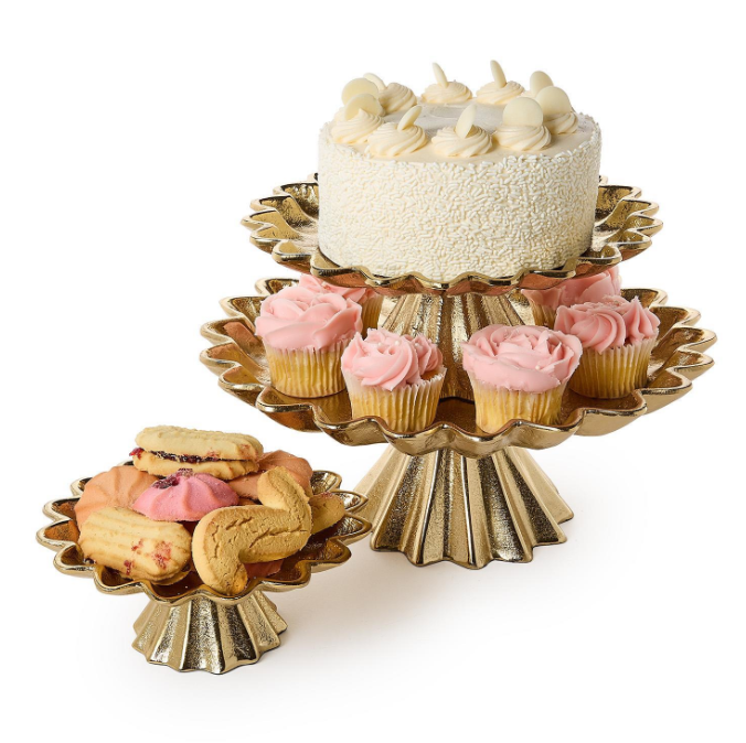 A three-tier gold pedestal stand with scalloped edges, shown holding a white frosted cake on the top tier, pink-frosted cupcakes on the middle tier, and assorted cookies on the bottom tier, against a plain white background.