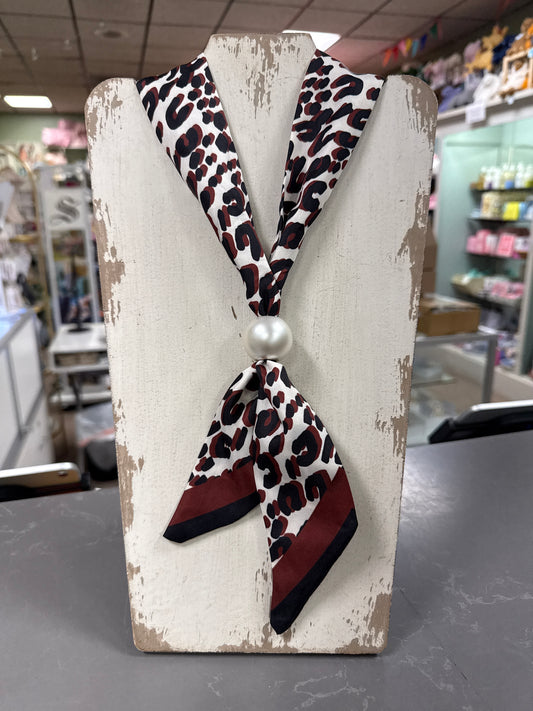 A leopard print neck scarf with a faux pearl center, displayed on a distressed white wooden stand inside a retail store.