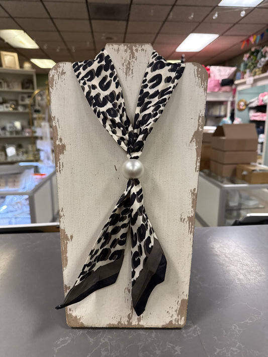 A black and white leopard print neck scarf tied in a knot with a white pearl center, displayed on a distressed white wooden stand inside a retail store.