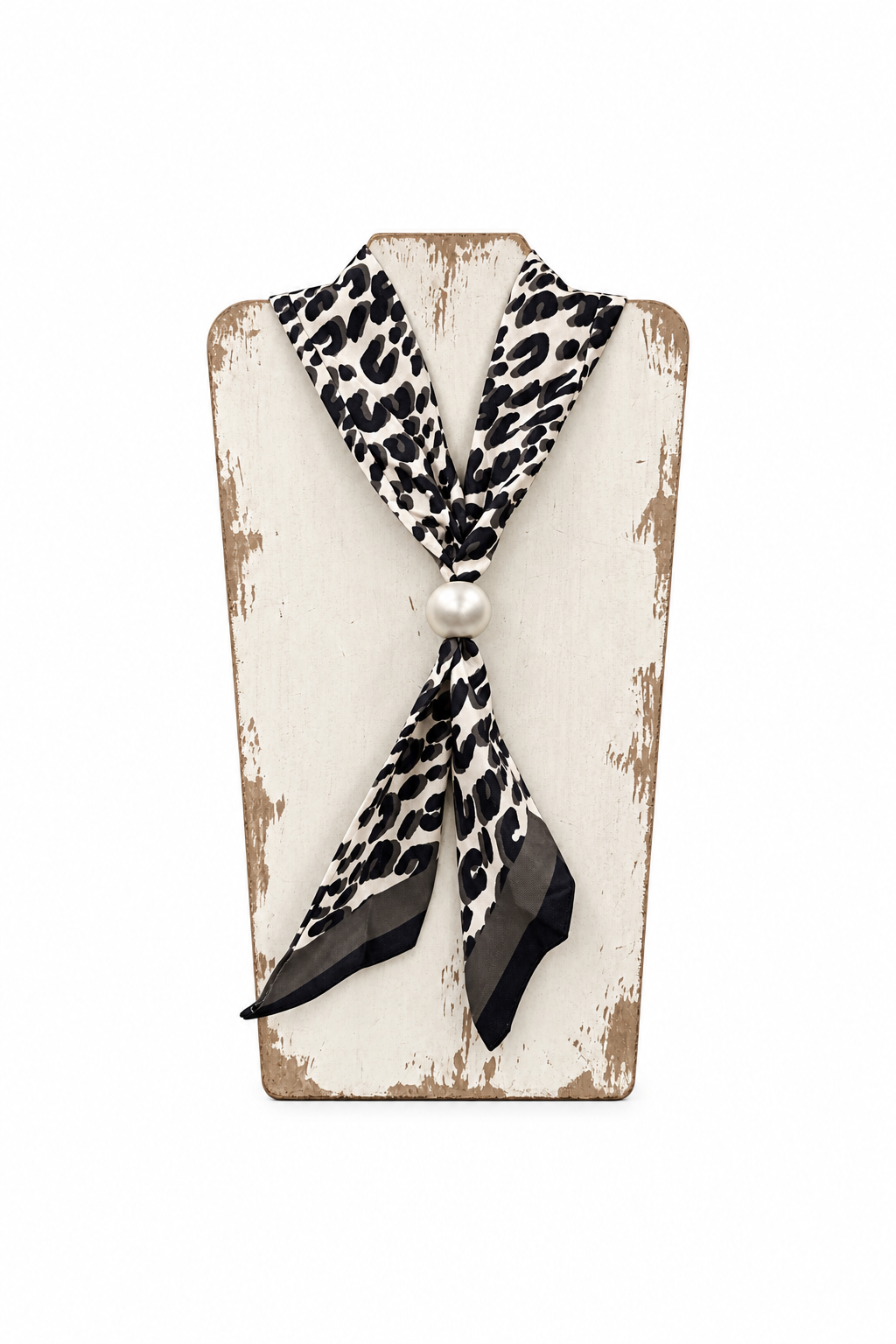 A black and white leopard print neck scarf tied in a knot with a white pearl center, displayed on a distressed white wooden stand inside a retail store.