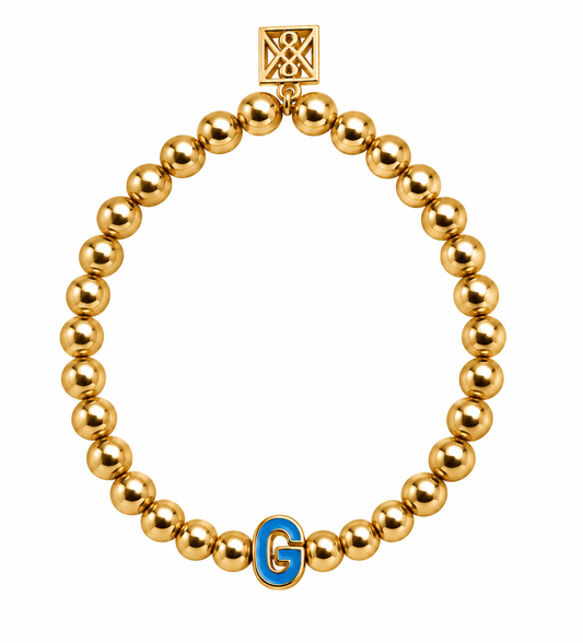 A gold beaded stretch bracelet featuring a square clasp with an interlocking X design and a blue and gold letter 'G' charm, shown on a plain white background.