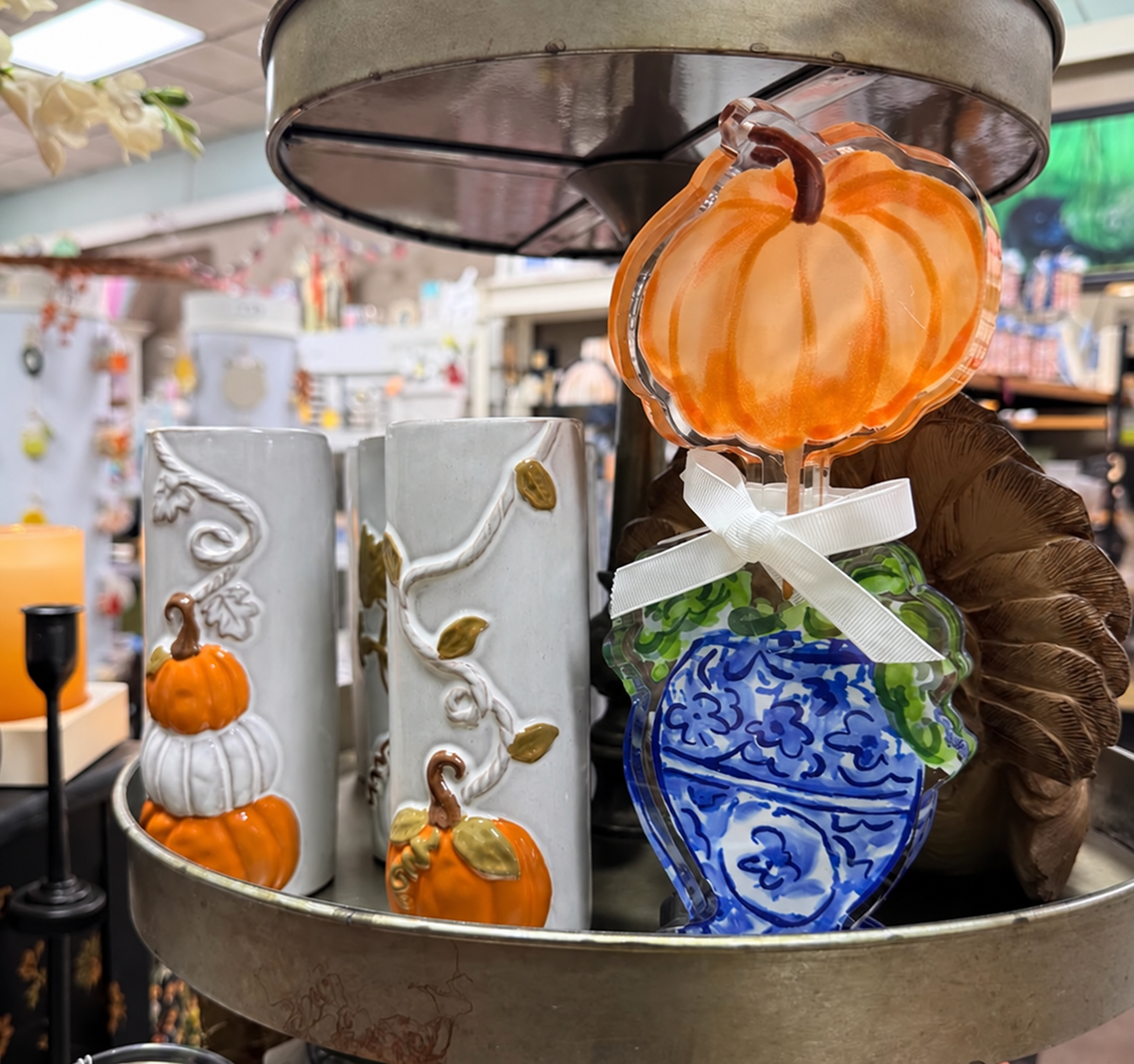 A decorative autumn-themed display on a metal tray, featuring two white ceramic candle holders with orange and white pumpkins and vine motifs, a blue and white porcelain-style ornament, a clear orange pumpkin topper, and a brown feather.