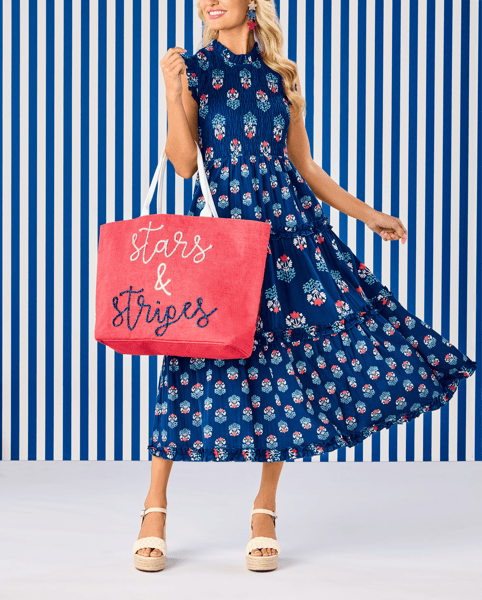 A woman models a navy blue sleeveless midi dress with a red, white, and blue floral pattern, holding a pink tote bag that reads 'stars & stripes' against a blue and white striped background.