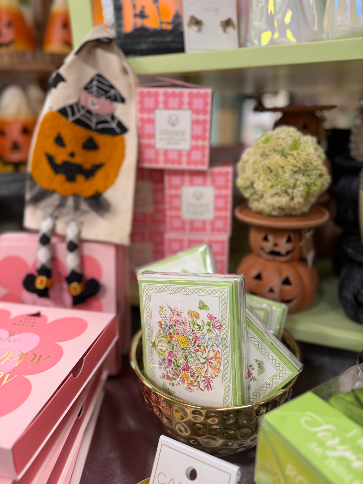 A close-up of a retail display featuring a stack of floral greeting cards in a gold bowl, surrounded by Halloween decorations including a jack-o'-lantern candle holder, a witch hat bag, and pink gift boxes.