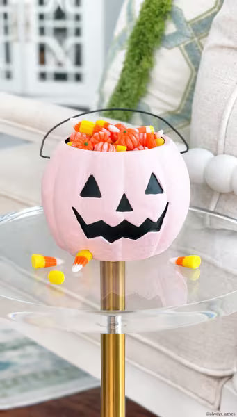 A pink jack-o'-lantern bucket filled with candy corn and orange pumpkins, displayed on a glass side table in a living room setting.