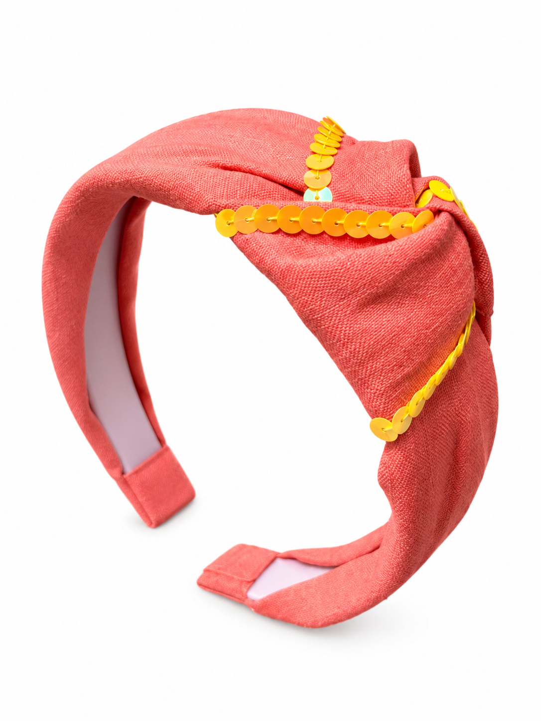 A wide fabric headband in a coral pink color, featuring a twisted knot design adorned with yellow sequins and trim, shown on a plain white background.