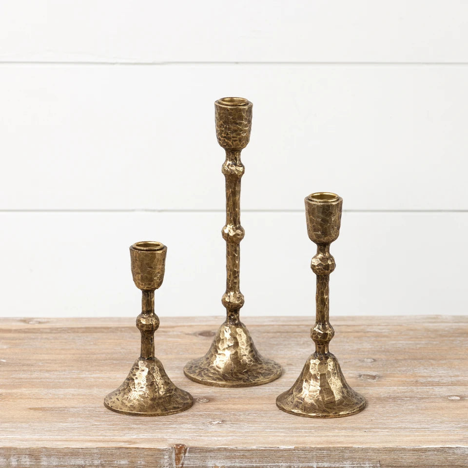 Three rustic gold-toned metal candlestick holders with cream taper candles, styled on a wooden surface against a white shiplap wall with holiday decor.