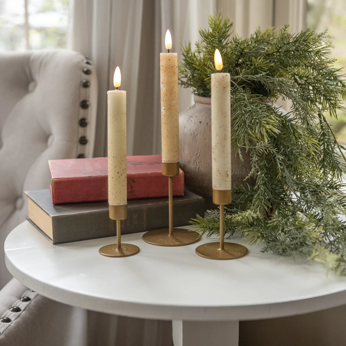 Three lit taper candles in gold holders arranged on a white table beside stacked books and greenery in a softly lit interior setting.