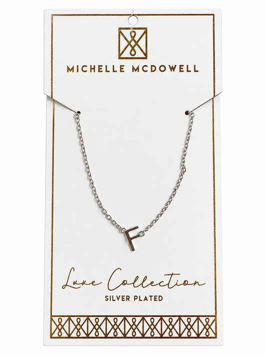 A silver-plated initial necklace with the letter 'F' pendant, displayed on a white Michelle McDowell brand card.