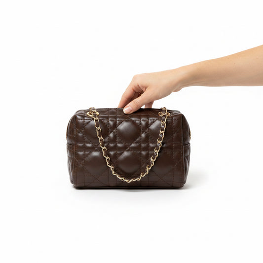 A dark brown quilted leather handbag with a gold chain strap, resting on a gray retail counter inside a store.