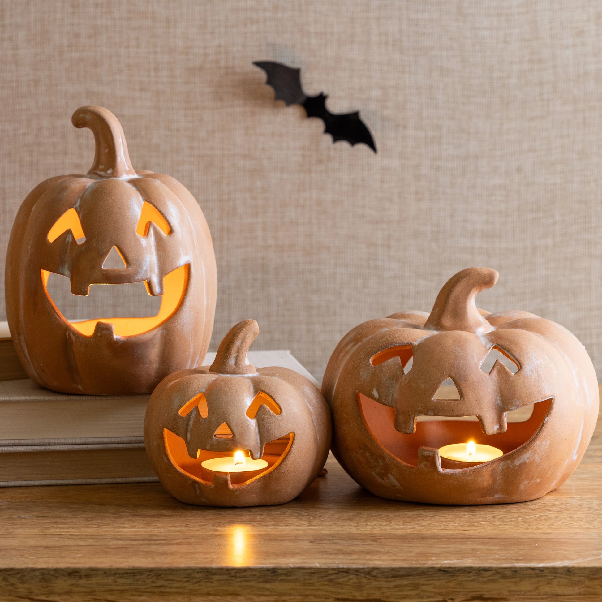 Three ceramic jack-o'-lanterns with glowing interiors on a wooden surface, with a bat decoration in the background.