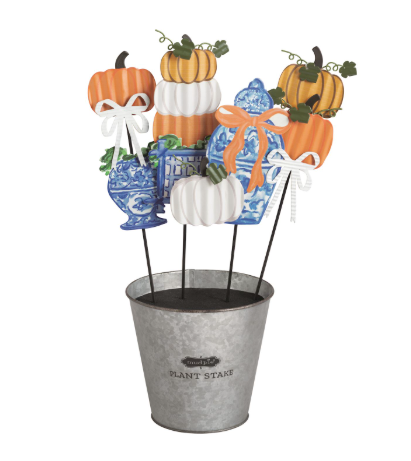 An autumn-themed decorative centerpiece featuring stacked pumpkins, blue-and-white porcelain-style vases, wrapped gift boxes with orange ribbons, and a galvanized metal bucket labeled 'Plant Stakes' on a plain white background.
